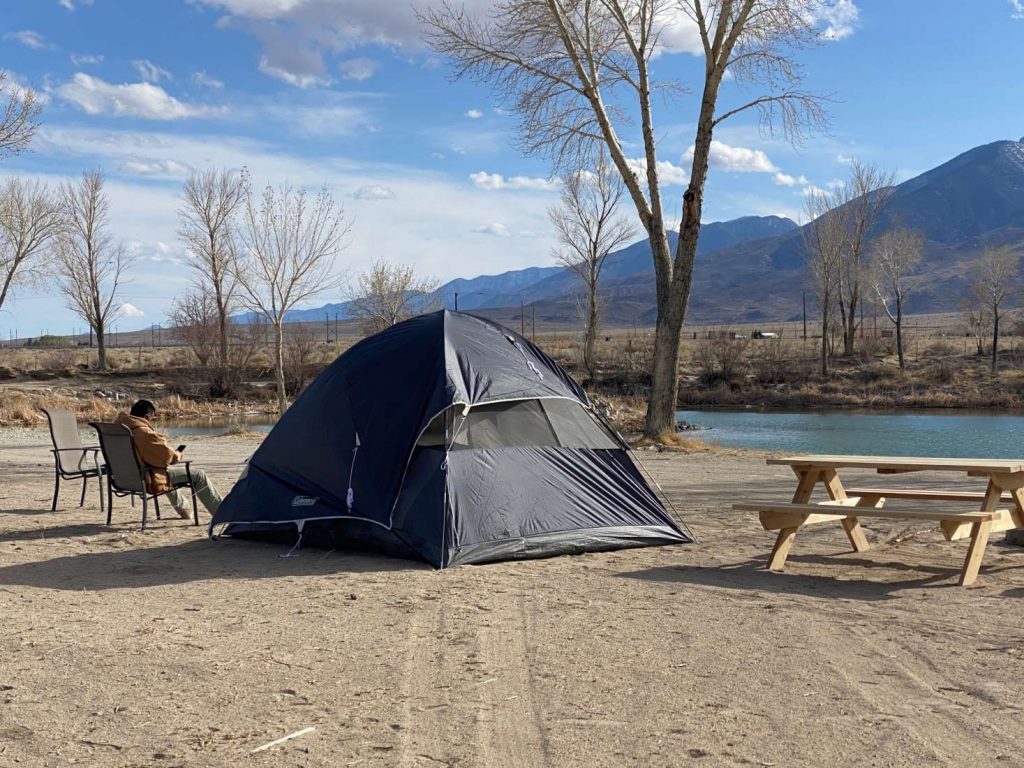 A person looking at his phone while sitting on a chair next to a tent by the lake in a sunny day.